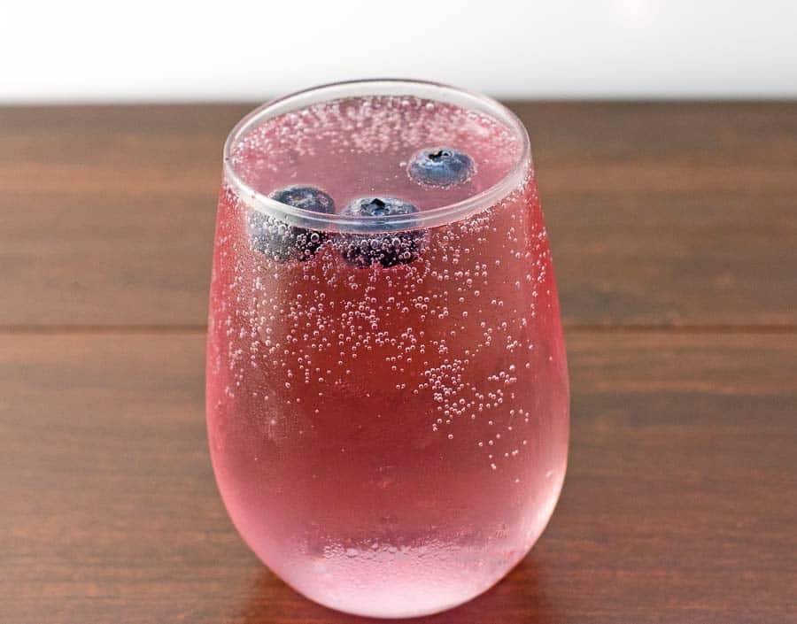 A finished mixed berry vodka spritzer in a clear stemless wine glass with frozen blueberries being used as ice