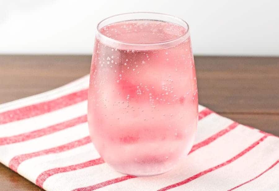 The finished mixed berry spritzer in a stemless wine glass sitting on a striped red and white towel that is on a wood  table with a marble background.