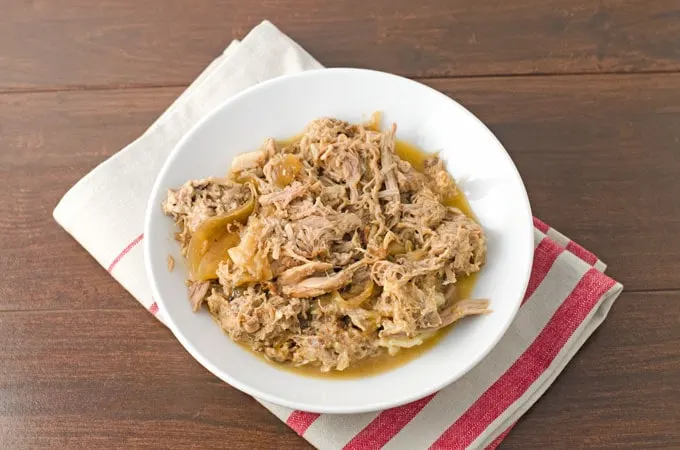 Cooked pulled pork and juices in a shallow white round bowl sitting on a red and white striped towel on a dark wood background.