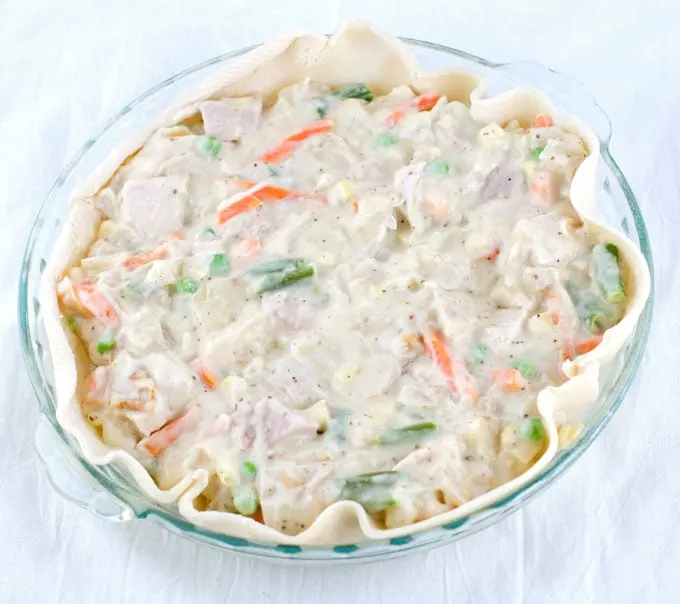 A whole unbaked chicken pot pie without the top crust in a glass pie pan with handles on a white cheesecloth background.
