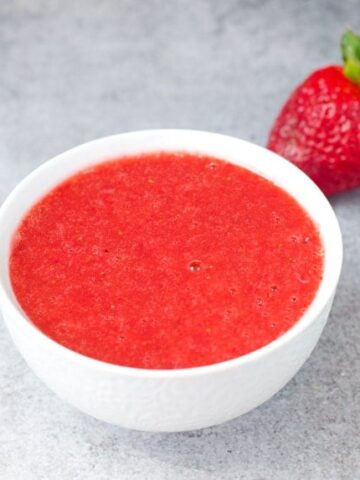 A white bowl of no-cook strawberry sauce with a fresh strawberry in the background