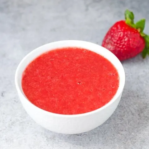 A white bowl of no-cook strawberry sauce with a fresh strawberry in the background
