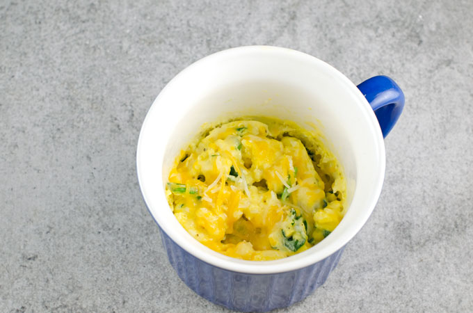 Cooked scrambled eggs in blue mug with white interior on a light gray background.