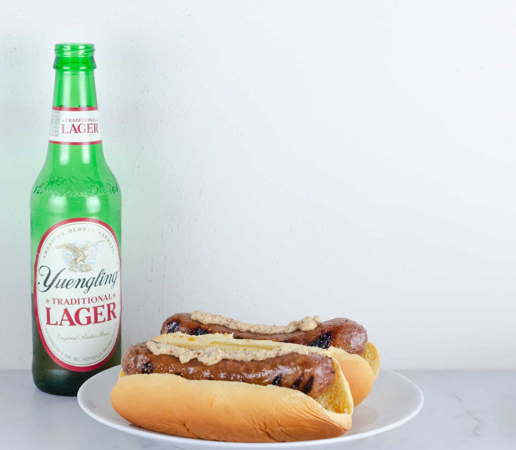 Honey Mustard Beer Glazed Bratwurst Recipe photo - brats in buns on a plate paired with yuengling lager in a bottle