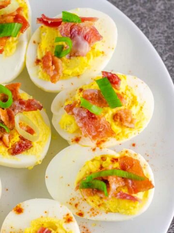 deviled eggs garnished with cayenne pepper, bacon, and sliced green onion on a white plate on a black slate countertop