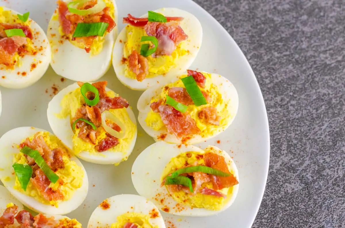 deviled eggs garnished with cayenne pepper, bacon, and sliced green onion on a white plate on a black slate countertop