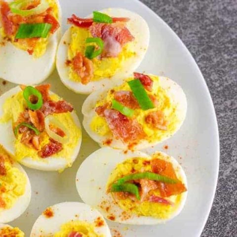 close up of bacon deviled eggs garnished with chopped bacon, cayenne powder, and sliced green onion on top of a white plate on a black slate surface