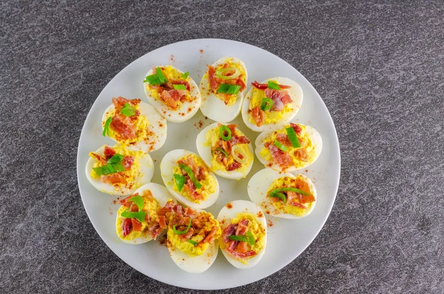 completed bacon deviled eggs garnished with cayenne, bacon, and slices of green onion on a white serving plate on top of a black slate counter top