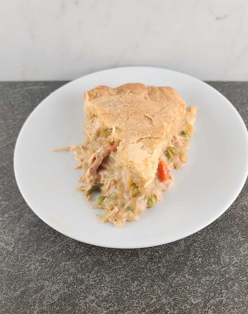An overhead shot of  a slice of baked chicken pot pie with the tip facing the front on a small white round plate on a slate surface and marble background.