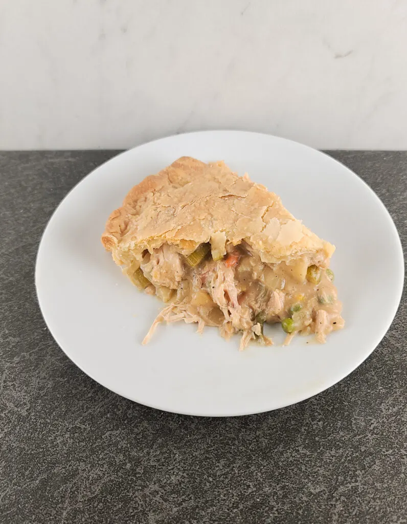 A overhead side view of a baked slice of chicken pot pie sitting on a white plate that's on a black slate surface with a marble background.