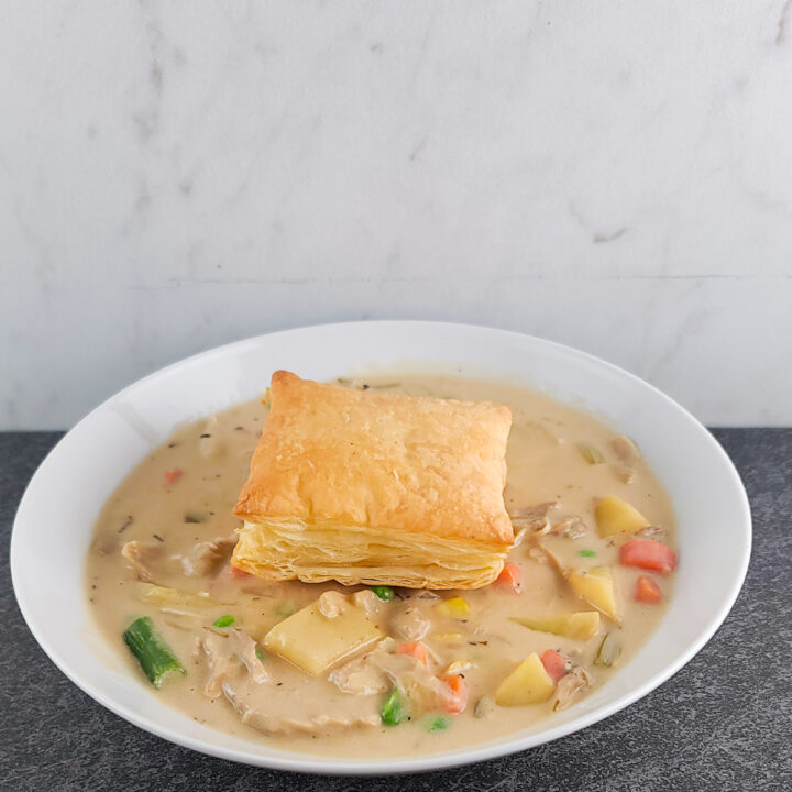 Chicken pot pie soup in a shallow white bowl with a square of baked puff pastry on top.