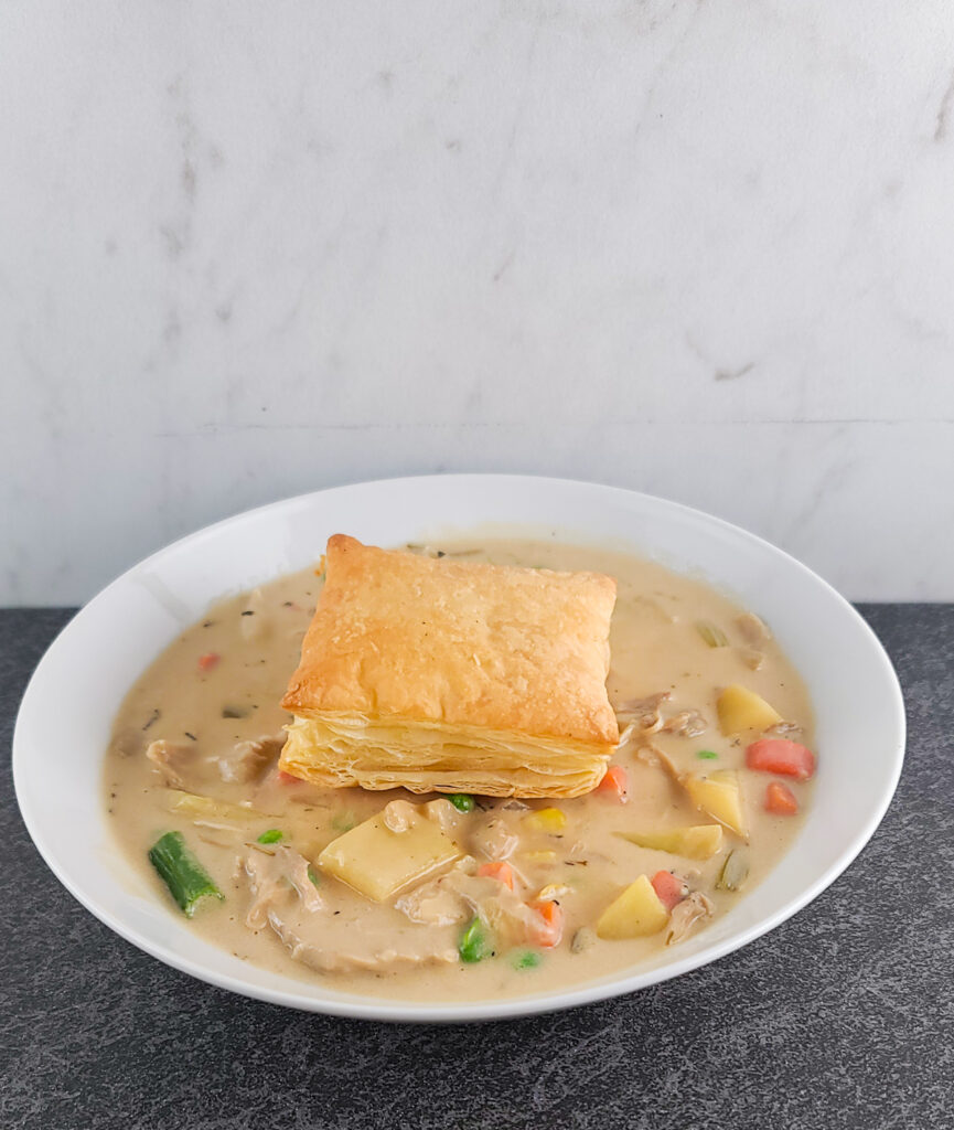 Chicken pot pie soup in a shallow white bowl with a square of baked puff pastry on top.