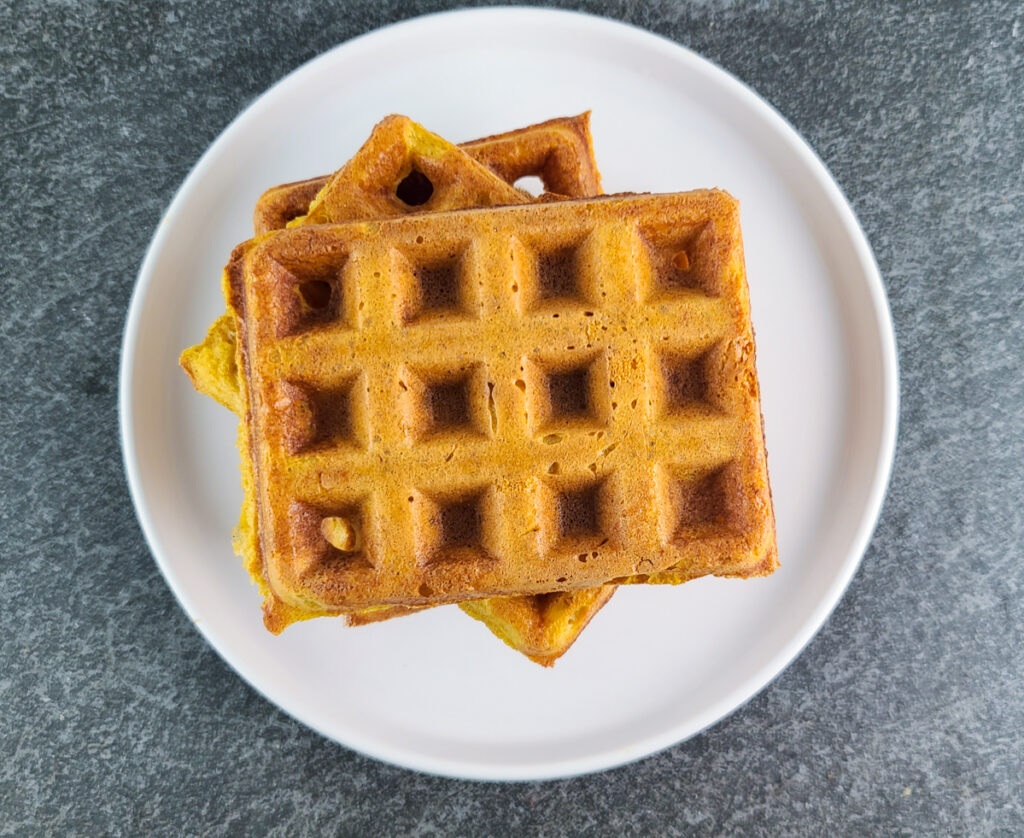overhead shot of 3 pumpkin waffles stacked on top of each other on a shallow white plate sitting on a slate surface