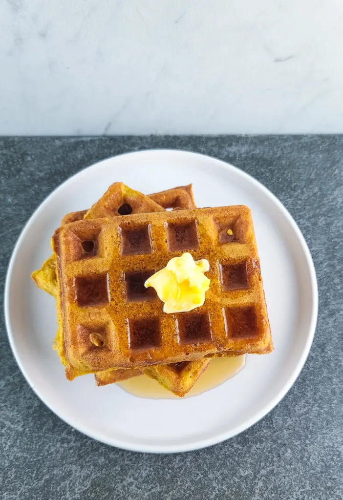 overhead shot of 3 pumpkin waffles stacked on top of each other with a pat of butter and heavy drizzle of maple syrup on a shallow round white plate on a slate surface with a marble background