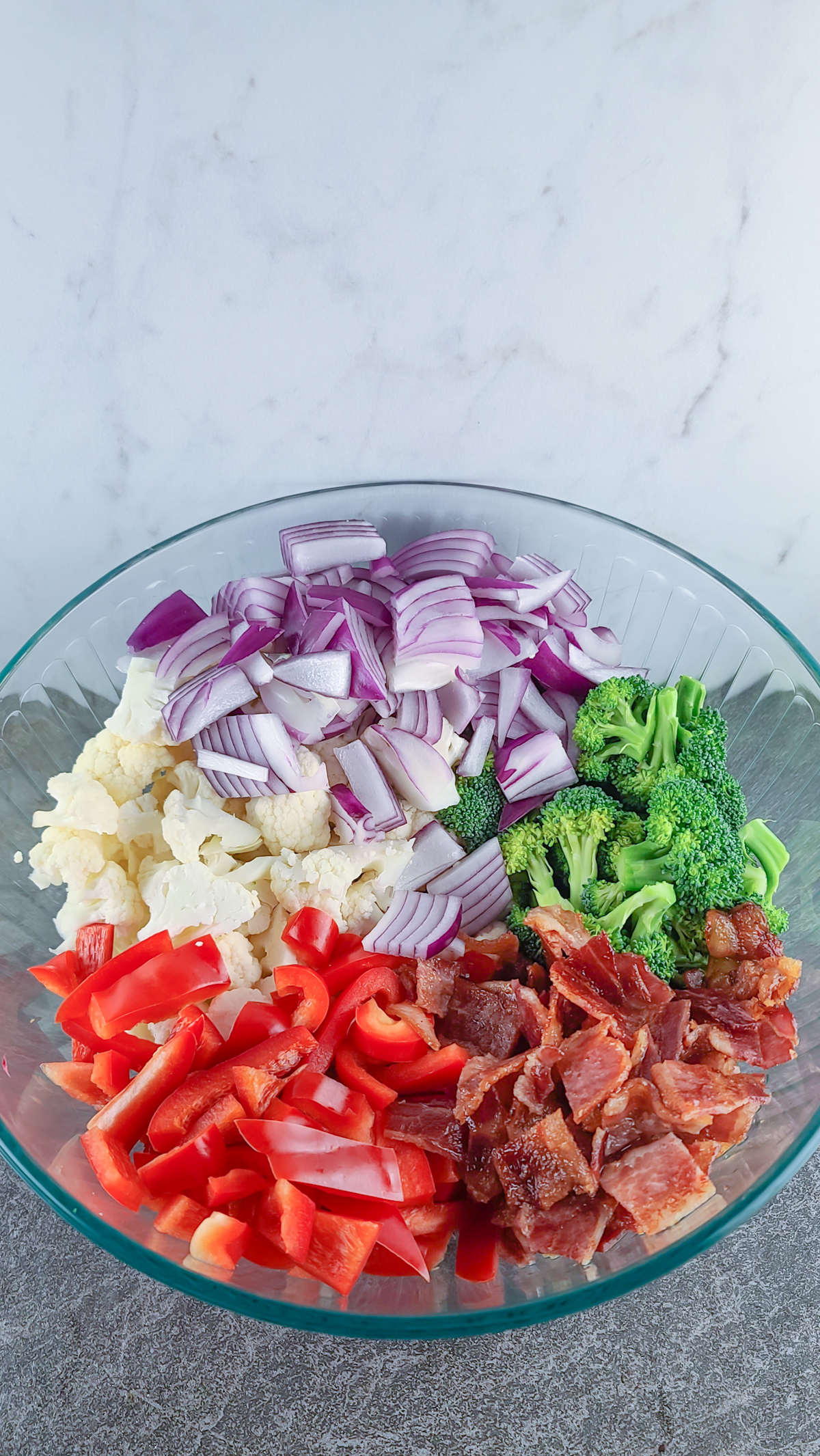 broccoli, cauliflower, red onion, red bell pepper, and bacon in a glass bowl on a slate surface with marble background