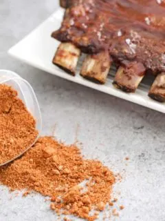 Plastic container on its side with pork dry rub flowing out of it. Prepared ribs using the rub in the background