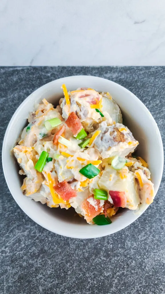 overhead closeup of a serving of the prepared baked potato salad in a round white bowl on a black slate background with marble backsplash