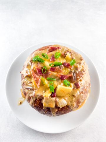 Angled shot of prepared bacon cheeseburger soup in a bread bowl on a white round plate.