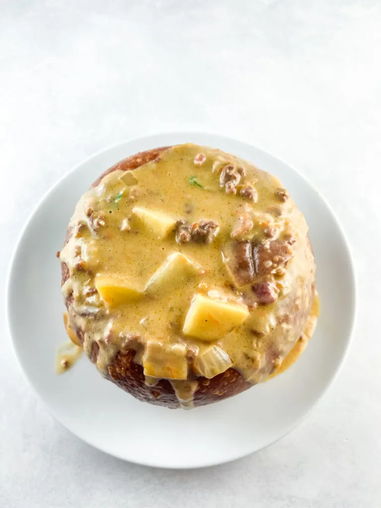 A bread bowl filled with cheeseburger soup flowing over the edges onto a round white plate.