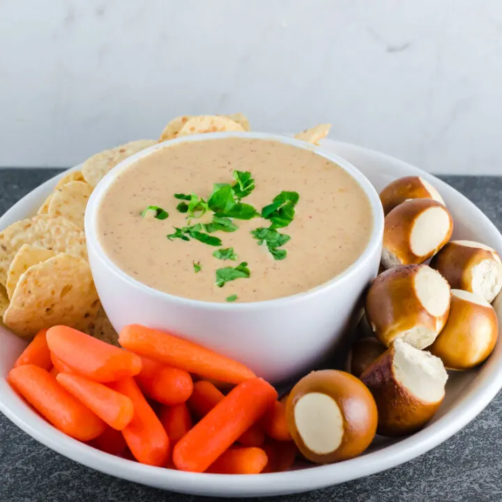 Queso dip in a round white bowl topped with chopped parsley nestled in a bigger bowl with pretzel bites, baby carrots, and tortilla chips for dipping.