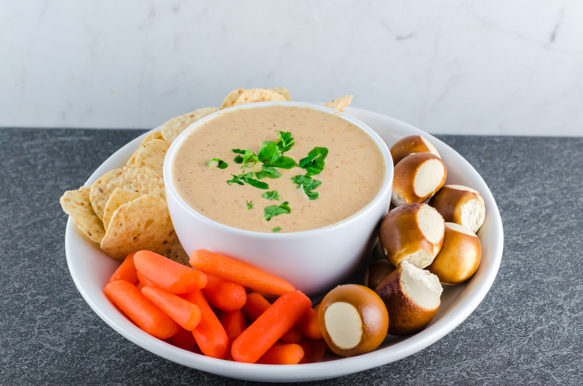 Queso dip in a round white bowl topped with chopped parsley nestled in a bigger bowl with pretzel bites, baby carrots, and tortilla chips for dipping.