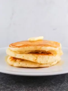 A stack of three buttermilk pancakes on a round white plate topped with a pat of butter and maple syrup.