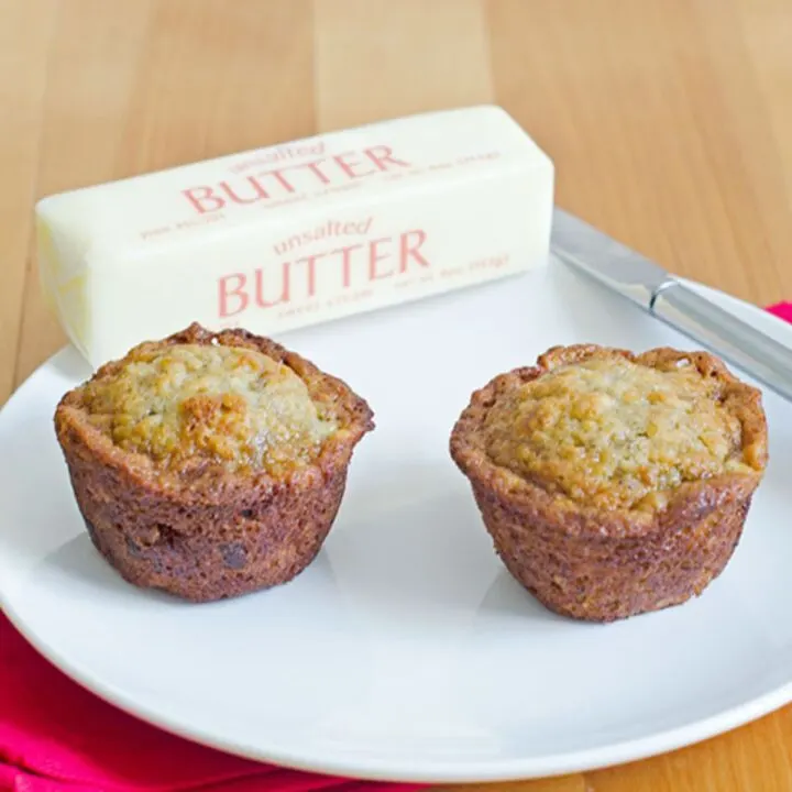 Two chocolate chip banana muffins on a small round white plate with a stick of butter and a dinner knife.