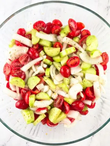 Ingredients for cucumber, tomato, and onion salad mixed together in a glass mixing bowl.