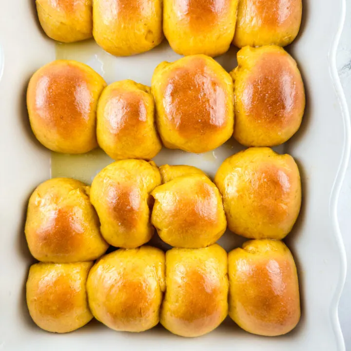 Baked pumpkin dinner rolls in a white baking dish that have been brushed with melted butter.